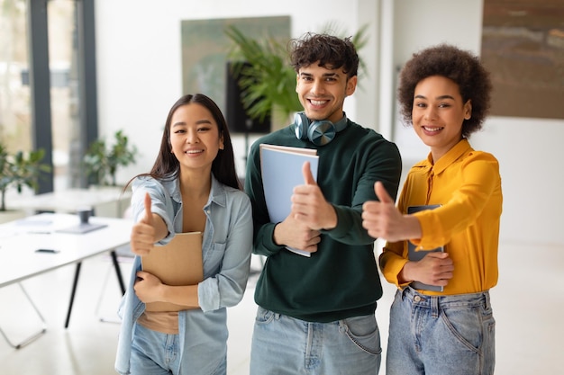 Felices estudiantes diversos gesticulando con el pulgar hacia arriba de pie en el espacio de coworking aprobando su universidad