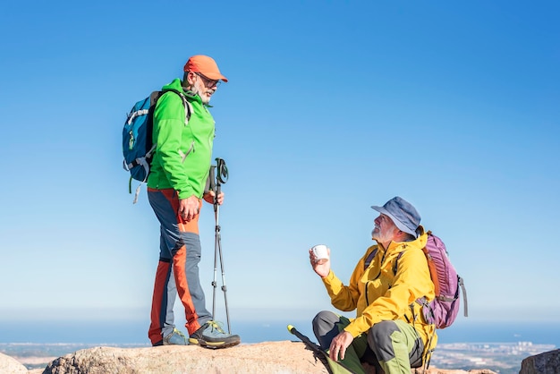 Felices excursionistas ancianos amigos con mochilas sentados hablando en la cumbre después de una caminata de montaña tomando un descanso