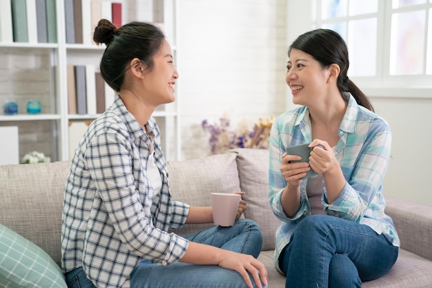 Felices hermanas asiáticas descansan en el sofá de la sala de estar hablando sobre la planificación de la vida futura sostienen tazas bebiendo té. dos chicas jóvenes mejores amigas en camisa y jeans charlando riéndose con una taza de café caliente en las manos.