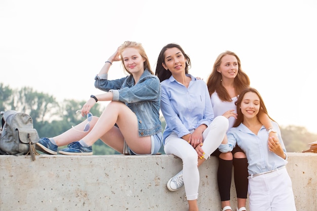 Felices y lindas amigas sentadas en el borde de hormigón y mirando la cámara