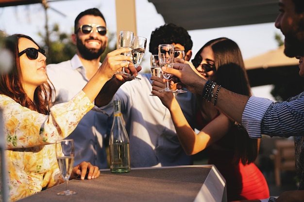 Felices mejores amigos en la fiesta de la terraza brindando con vino blanco Prosecco en una tarde de verano jóvenes tocando flautas y divirtiéndose gente de moda despreocupada el fin de semana en la fiesta de la azotea