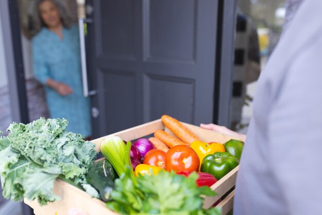 Foto felices mujeres caucásicas mayores abriendo la puerta de la casa y dando la bienvenida al mensajero con verduras. compras de comestibles en línea.