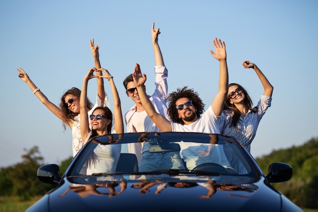 Felices las niñas y los chicos con gafas de sol están sentados en un descapotable negro en la carretera levantando las manos y sonriendo en un día soleado. .