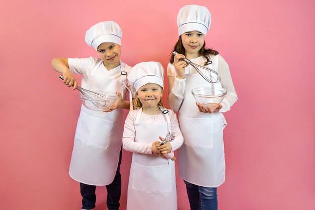 Felices niños chef con gorras y delantales, sosteniendo batidores y tazas sobre un fondo rosa, afiche, postal.