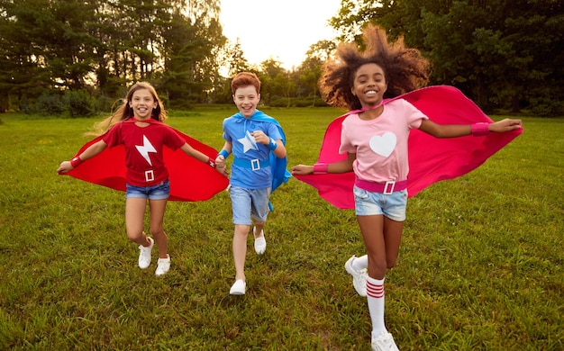 Felices los niños diversos en trajes de superhéroe sonriendo y corriendo sobre césped mientras juegan juntos en el parque al atardecer