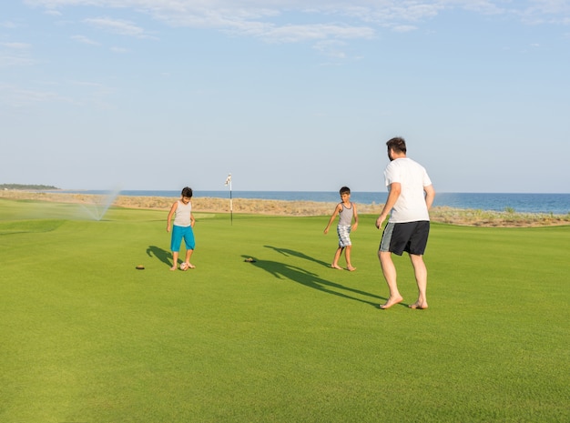 Felices los niños y el padre en terreno de hierba de golfo de verano que se divierten y tiempo feliz jugando al fútbol