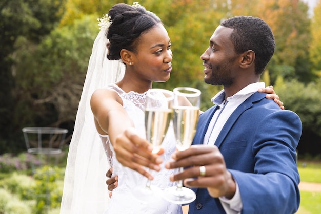 Felices novia y novio afroamericanos brindando con champán sonriéndose el uno al otro en un jardín soleado