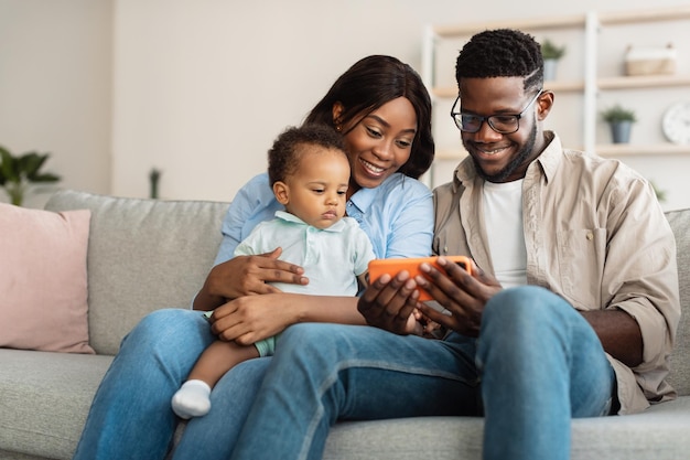 Felices padres afroamericanos jóvenes que muestran una aplicación educativa divertida en el teléfono móvil a un hijo pequeño y lindo, relajándose juntos en el sofá, familia emocionada usando un dispositivo digital, viendo videos