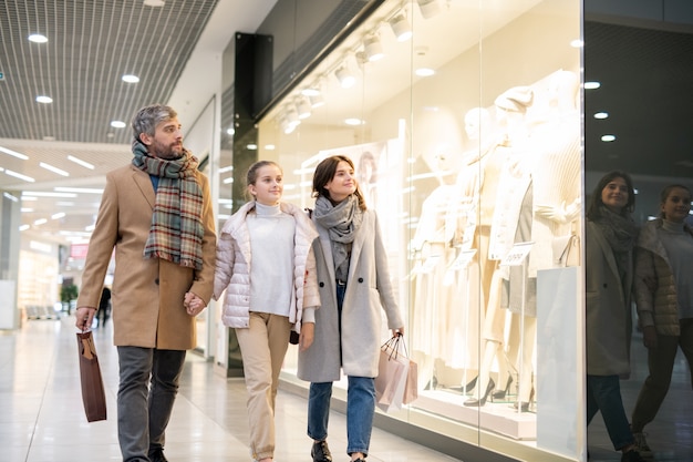 Felices padres e hija mirando la nueva colección de ropa casual en el escaparate mientras se relajan en el centro comercial durante la venta de temporada