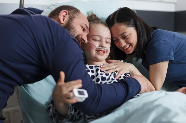 Niña enferma feliz abrazada por padres alegres y sonrientes en la sala de  pediatría del hospital. madre y padre alegres abrazando a una hija enferma  sentada en la cama de un paciente