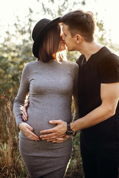 Foto felices padres jóvenes mamá y papá abrazando topetón de bebé besándose disfrutando de un hermoso momento al atardecer concepto de paternidad elegante pareja embarazada cogidos de la mano en el vientre a la luz del sol en el parque de otoño