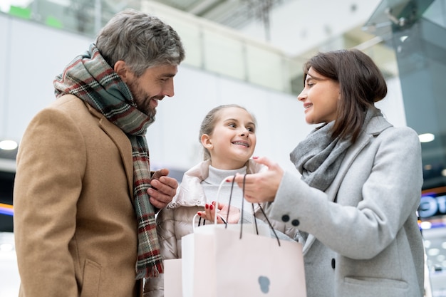 Felices padres jóvenes mirando a su hija mientras la madre muestra una nueva compra en una bolsa de papel después de comprar