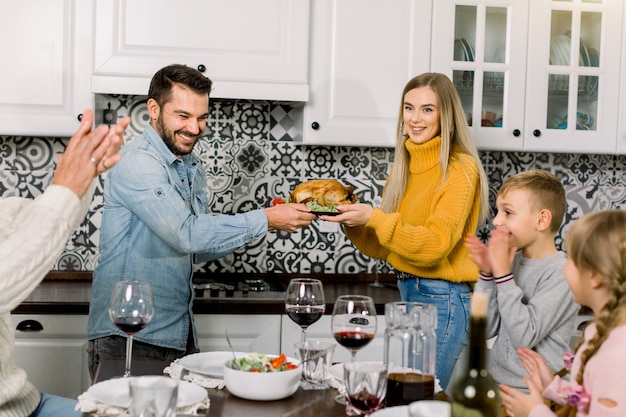 Felices padres jóvenes, mujer y hombre, ofreciendo pollo asado a familiares, niños y abuelo, cenando en casa