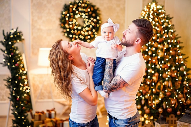 Felices padres jóvenes parados cerca del árbol de Navidad en la sala de estar mientras sostienen a una niñita agradable