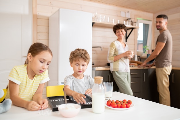 Felices padres jóvenes en ropa casual discutiendo planes para el día mientras están de pie en la cocina después del desayuno y miran a sus hijos jugando