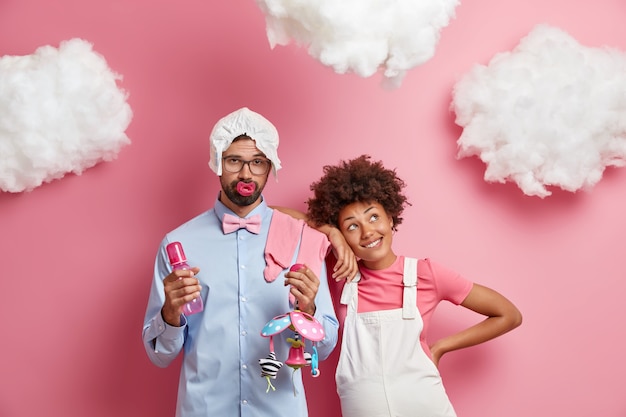 Felices padres de raza mixta esperan que el bebé se prepare para convertirse en padre y madre. Feliz mujer embarazada afroamericana se inclina sobre el hombro del marido que sostiene el biberón y el juguete. Concepto de expectativa