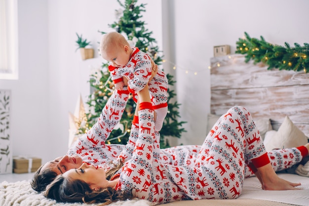 Felices padres con su pequeña hija en ropa de vacaciones con ciervos impresos y copos de nieve divirtiéndose en la cama en una acogedora habitación con un árbol de Navidad