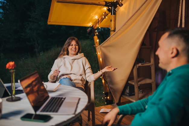 Felices parejas familiares independientes descansan después de trabajar con una laptop en una acogedora tienda glamping en las noches de verano Tienda de campaña de lujo para vacaciones al aire libre y vacaciones Concepto de estilo de vida