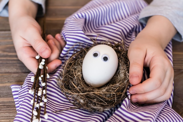 Felices Pascuas. Huevo sin pintar con ojos en un nido de pájaros, manos de niños y ramas de sauce en una mesa de madera.