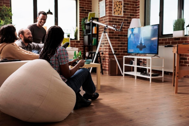 Felices y sonrientes amigos diversos sentados en un sofá en la sala de estar mientras juegan videojuegos en la consola. La gente en casa se relaja con juegos en un dispositivo de entretenimiento mientras disfruta de bocadillos y bebidas.