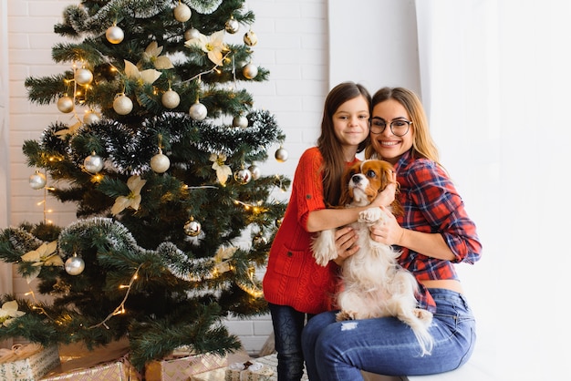 ¡Felices vacaciones de invierno! Mujer morena positiva abraza pose de niña con regalos en el piso en la habitación, perro cerca, divertirse cerca del árbol de Navidad.