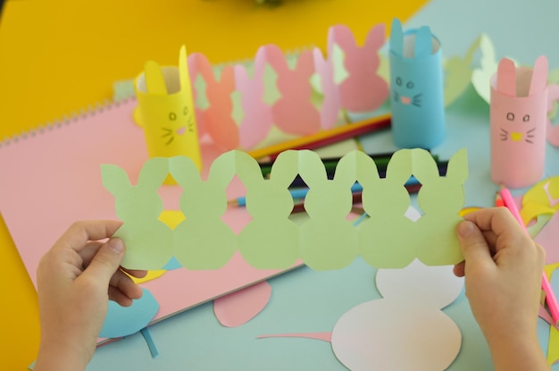 Felices vacaciones de Pascua preparándose para la sala de decoración de Pascua Primer plano de las manos de los niños desplegando una guirnalda verde de bricolaje de conejitos de Pascua hechos de papel de colores Celebración cristiana tradiciones familiares