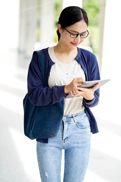 Felicidad alegre joven estudiante asiática en ropa casual lectura y hacer una nota corta en la tableta para el examen en fondo borroso concepto de la escuela asiática