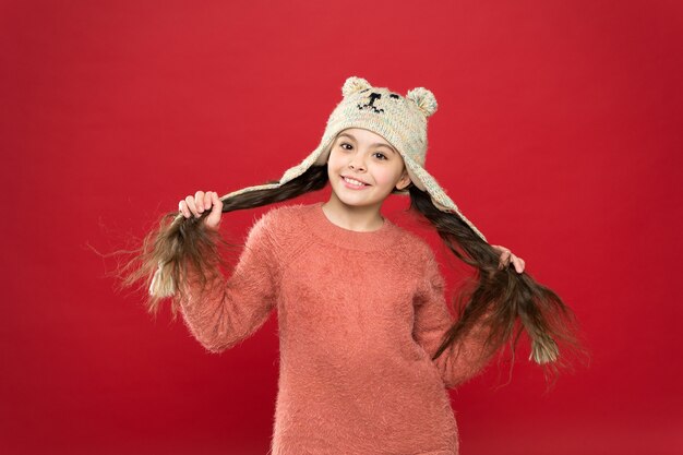 Felicidad y alegría. Traje de invierno. Fondo rojo del sombrero del desgaste del pelo largo del niño pequeño. Modelo lindo disfruta del estilo de invierno. Adorable osito. Niño pequeño usa gorro de punto. Accesorio de moda de invierno de niña.