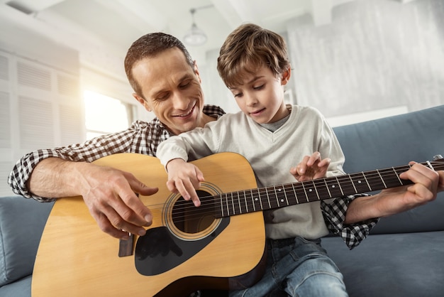 Felicidad. Atractivo niño rubio concentrado aprendiendo a tocar la guitarra mientras está sentado en el sofá y su padre sonriendo