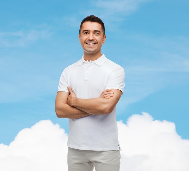 felicidad y concepto de la gente - hombre sonriente en camiseta blanca con los brazos cruzados sobre el cielo azul y el fondo de la nube