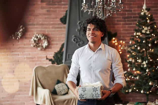 La felicidad está en las pequeñas cosas. Joven de pie en la hermosa habitación decorada y sosteniendo una caja de regalo blanca en la época de año nuevo.