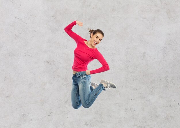 Foto felicidad, libertad, poder, movimiento y concepto de la gente - mujer joven sonriente saltando en el aire con el puño levantado sobre fondo de pared de hormigón gris