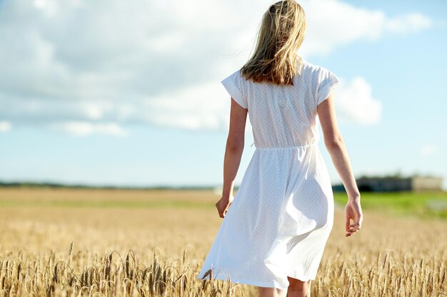 felicidad, naturaleza, vacaciones de verano, vacaciones y conceptos de personas - mujer joven en vestido blanco caminando por el campo de cereales