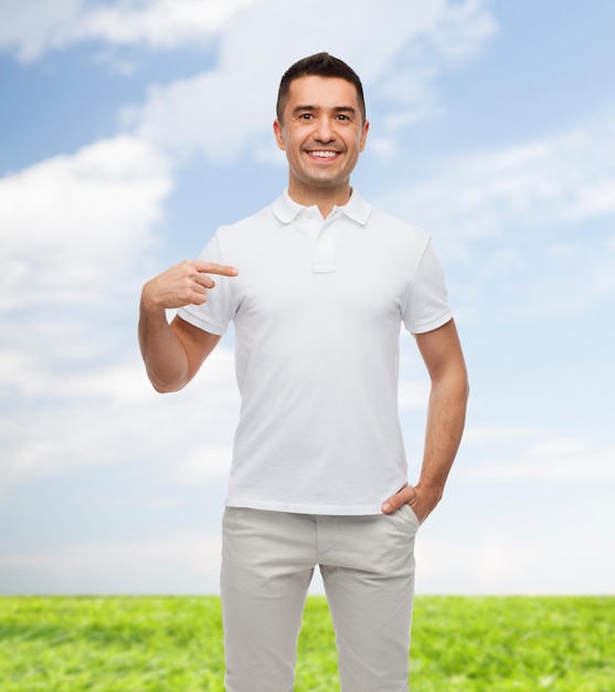 felicidad, publicidad, moda, gesto y concepto de la gente - hombre sonriente en camiseta apuntándose con el dedo sobre el cielo azul y el fondo de la hierba