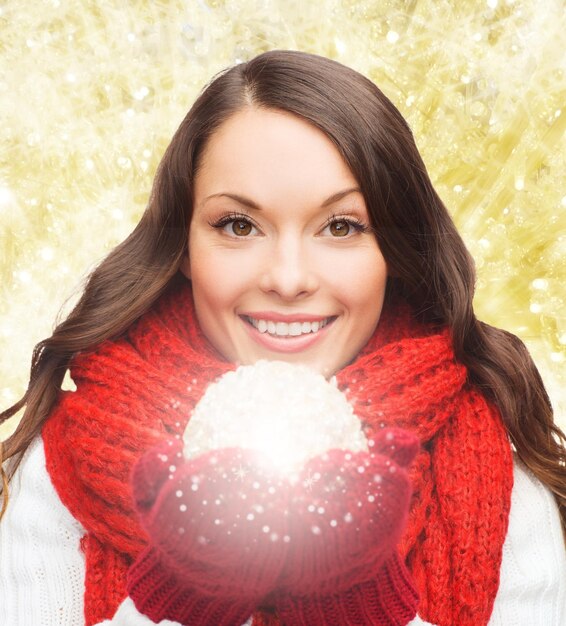 felicidad, vacaciones de invierno y concepto de la gente - mujer joven sonriente con bufanda roja y mitones con bola de navidad sobre fondo de luces amarillas