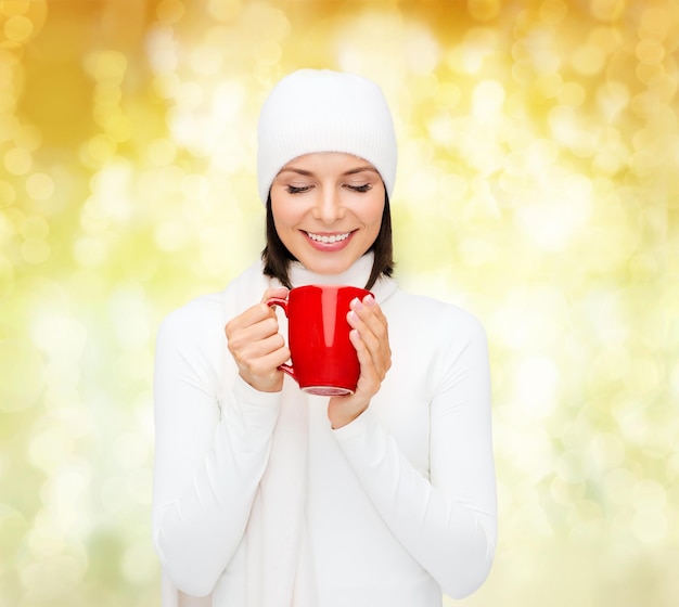 felicidad, vacaciones de invierno, navidad, bebidas y concepto de personas - mujer joven sonriente con ropa blanca cálida con taza roja sobre fondo de luces amarillas