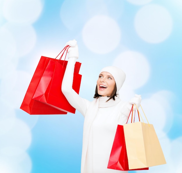 felicidad, vacaciones de invierno, navidad y concepto de la gente - mujer joven sonriente con sombrero blanco y mitones con bolsas de compras rojas sobre fondo de luces azules