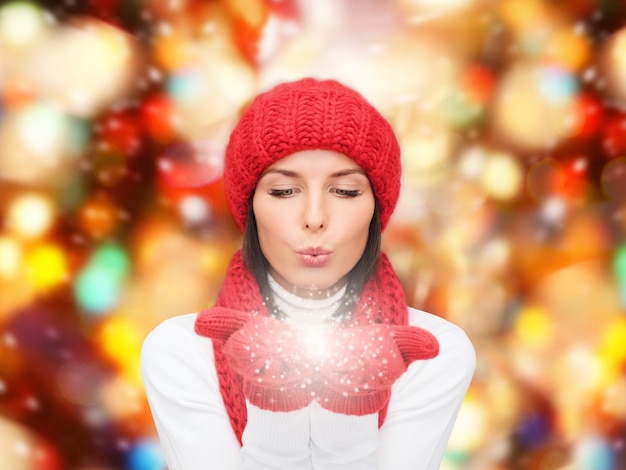 felicidad, vacaciones de invierno, navidad y concepto de la gente - mujer joven sonriente con sombrero rojo, bufanda y mitones sobre fondo de luces