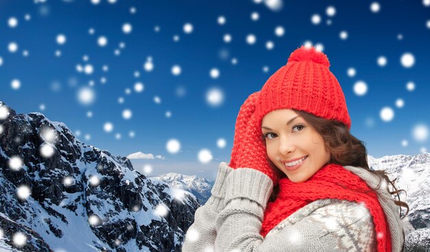 felicidad, vacaciones de invierno, navidad y concepto de la gente - mujer joven sonriente con sombrero rojo, bufanda y mitones sobre fondo de montañas nevadas