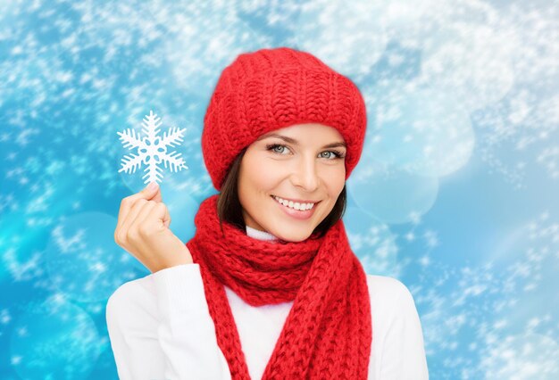 felicidad, vacaciones de invierno, navidad y concepto de la gente - mujer joven sonriente con sombrero rojo, bufanda y mitones sosteniendo copos de nieve sobre fondo de luces azules