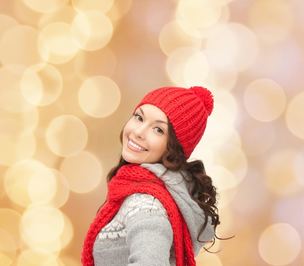 felicidad, vacaciones de invierno, navidad y concepto de la gente - mujer joven sonriente con sombrero rojo y bufanda sobre fondo de luces beige