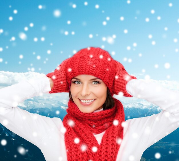 felicidad, vacaciones de invierno, turismo, viajes y concepto de personas - mujer joven sonriente con sombrero rojo y mitones sobre fondo de montañas nevadas