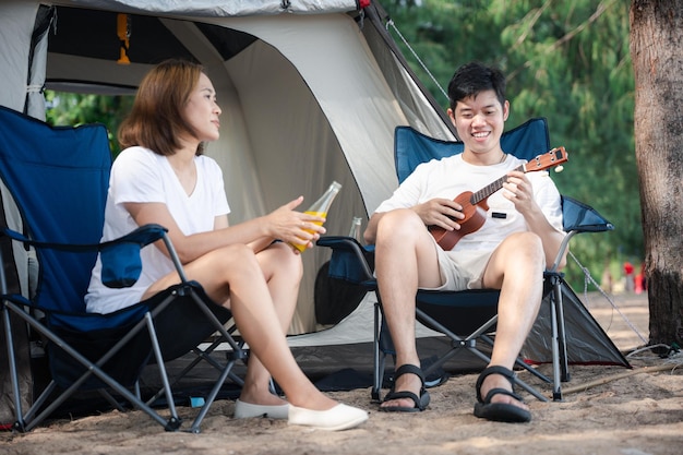 Felicidade de acampamento Um casal asiático alegre em sua tenda canta serenadas um ao outro com músicas de ukulele espalhando amor e sorrisos sob o pôr-do-sol A união é a sua melodia Acampamento ao ar livre
