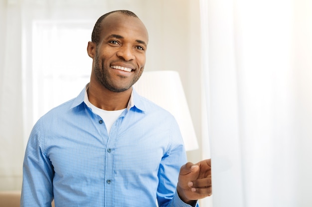 Felicidade dentro de mim. Homem afro-americano bem parecido e feliz, de pé, olhando pela janela e sorrindo