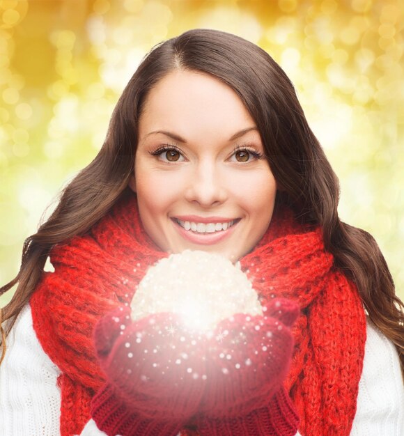 felicidade, férias de inverno e conceito de pessoas - jovem sorridente no lenço vermelho e luvas com bola de natal sobre fundo de luzes amarelas