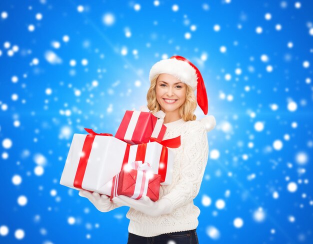 Felicidade, férias de inverno, natal e conceito de pessoas - jovem sorridente com chapéu de ajudante de papai noel com presentes sobre fundo azul nevando