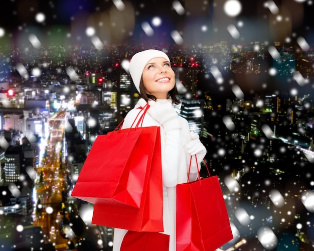 felicidade, férias de inverno, natal e conceito de pessoas - jovem sorridente de chapéu branco e luvas com sacolas de compras vermelhas sobre o fundo da noite da cidade nevada