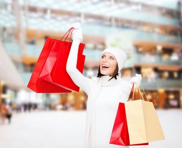 felicidade, férias de inverno, natal e conceito de pessoas - jovem sorridente de chapéu branco e luvas com sacos de compras vermelhos sobre fundo de centro comercial