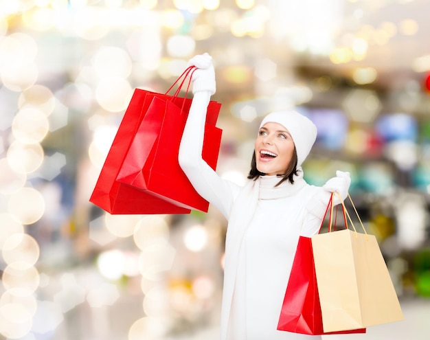 Felicidade, férias de inverno, natal e conceito de pessoas - jovem sorridente de chapéu branco e luvas com sacos de compras vermelhos sobre fundo de luzes