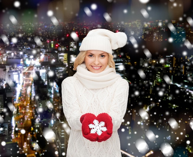 felicidade, férias de inverno, natal e conceito de pessoas - jovem sorridente de chapéu, cachecol e luvas segurando a decoração do floco de neve sobre o fundo da cidade de noite de neve